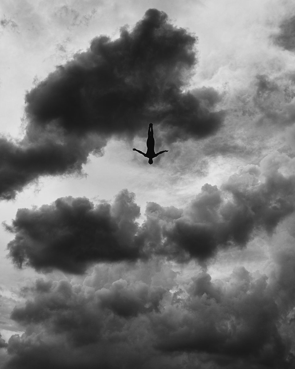 Monochrome image of a person in freefall against a backdrop of dramatic, swirling clouds, creating a striking sense of scale and freedom.
