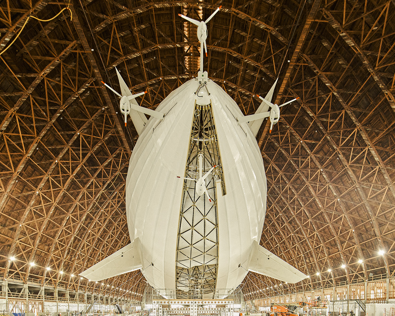 Large white dirigible with propellers hanging in a cavernous wooden hangar with lattice-work support beams, viewed from a low angle.
