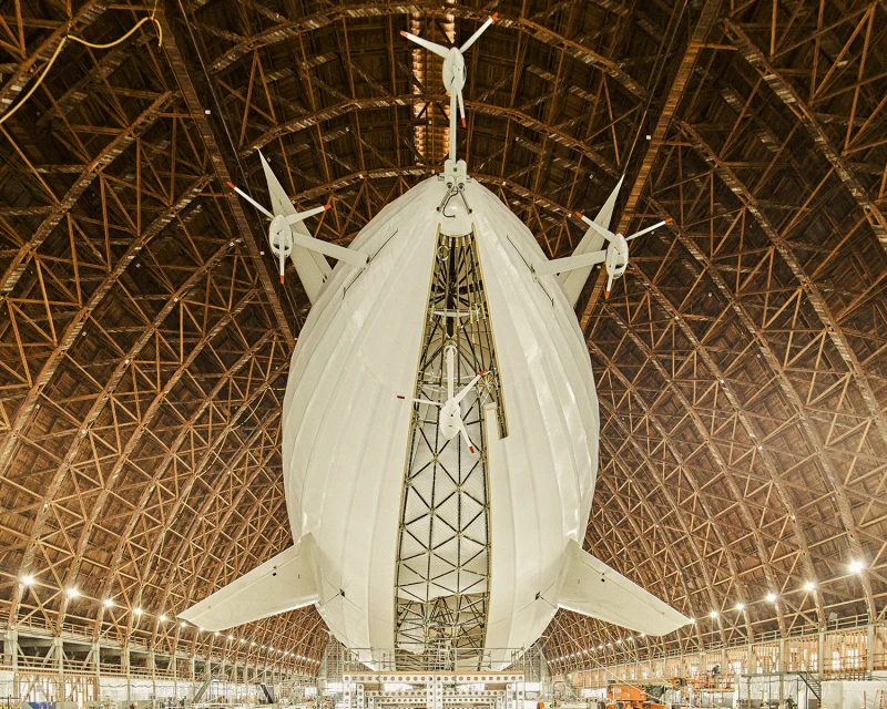 Large white dirigible with propellers hanging in a cavernous wooden hangar with lattice-work support beams, viewed from a low angle.