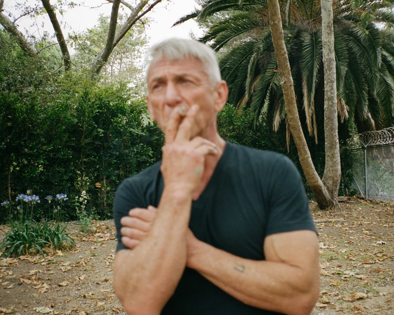 Mature adult male with gray hair and a dark shirt stands in a park-like setting with palm trees and green foliage, holding a cigarette.