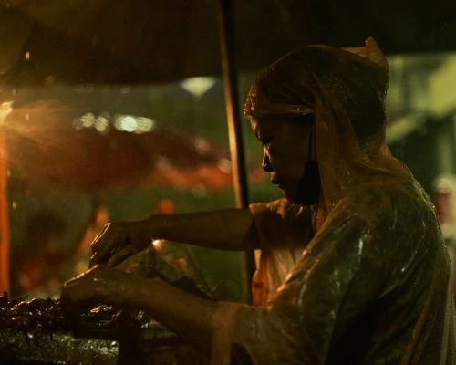 An individual in a translucent raincoat tending to an outdoor food stall, illuminated by nearby lighting, during what appears to be nighttime.