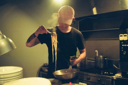 Chef holds steaming noodles in a restaurant kitchen, featuring stainless steel appliances, cookware, and professional cooking equipment.