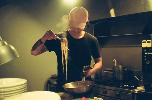Chef holds steaming noodles in a restaurant kitchen, featuring stainless steel appliances, cookware, and professional cooking equipment.