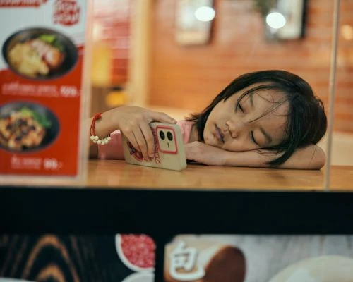 Young Asian girl asleep at a table with a smartphone next to her face.  Restaurant setting with muted colors and shallow focus.