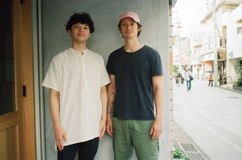 Two young adult men stand in front of a building with an alleyway street scene in the background, wearing casual attire in daylight.