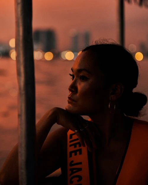 Portrait of a woman wearing a life jacket, resting her chin on her hand, with blurred city lights visible in the background at dusk.
