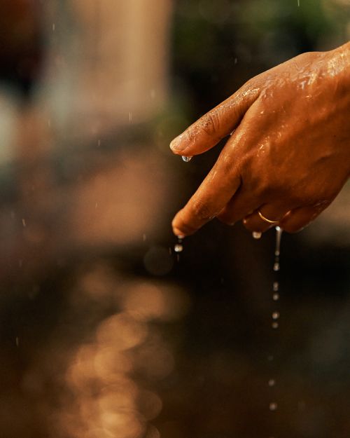 Close-up of an outstretched tan hand with water droplets falling from the index finger, set against a blurry brown background.