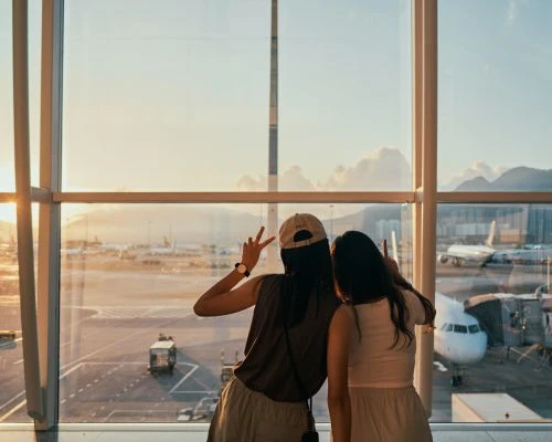 Two women at an airport pose looking out the window at airplanes and mountains in the distance during golden hour, travel photography.