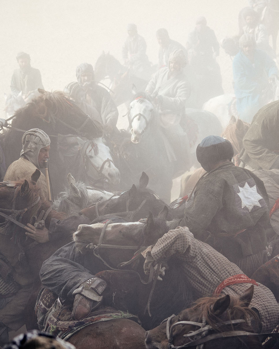 A group of male buzkashi riders on horseback compete in a cloud of dust, wearing traditional clothing and hats. The game takes place outdoors.