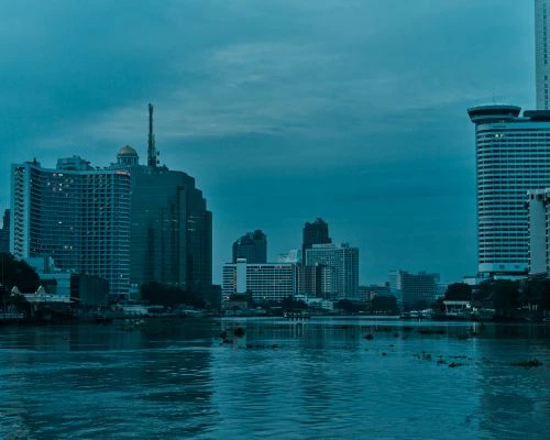 Cityscape at dusk featuring modern buildings and a wide river, under a blue-tinted sky, reflecting urban lights in the water.