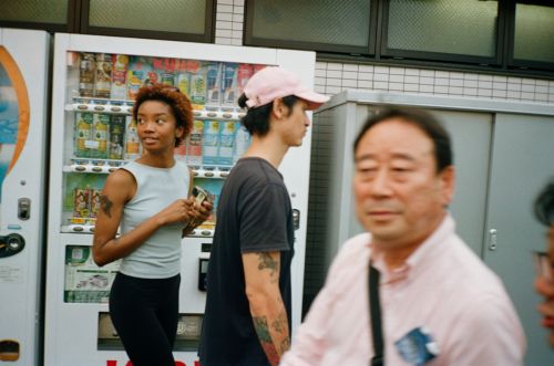 Three people stand near a vending machine in an urban setting. A woman with curly hair is looking to the left, holding an item.