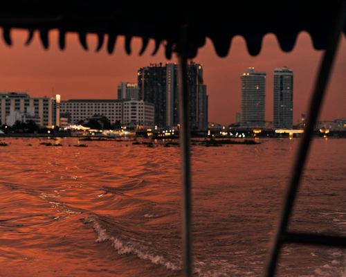 View of Bangkok city buildings and river at sunset from a boat. Water waves reflect the orange sky and city lights in the background.