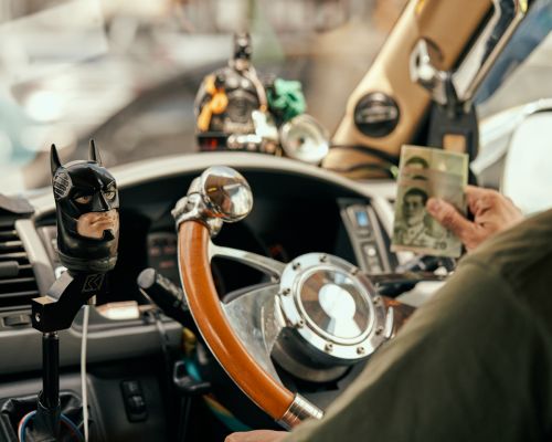 Car interior featuring a driver holding Thai Baht currency, a Batman shift knob, and decorative figurines on the dashboard.