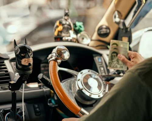 Car interior featuring a driver holding Thai Baht currency, a Batman shift knob, and decorative figurines on the dashboard.