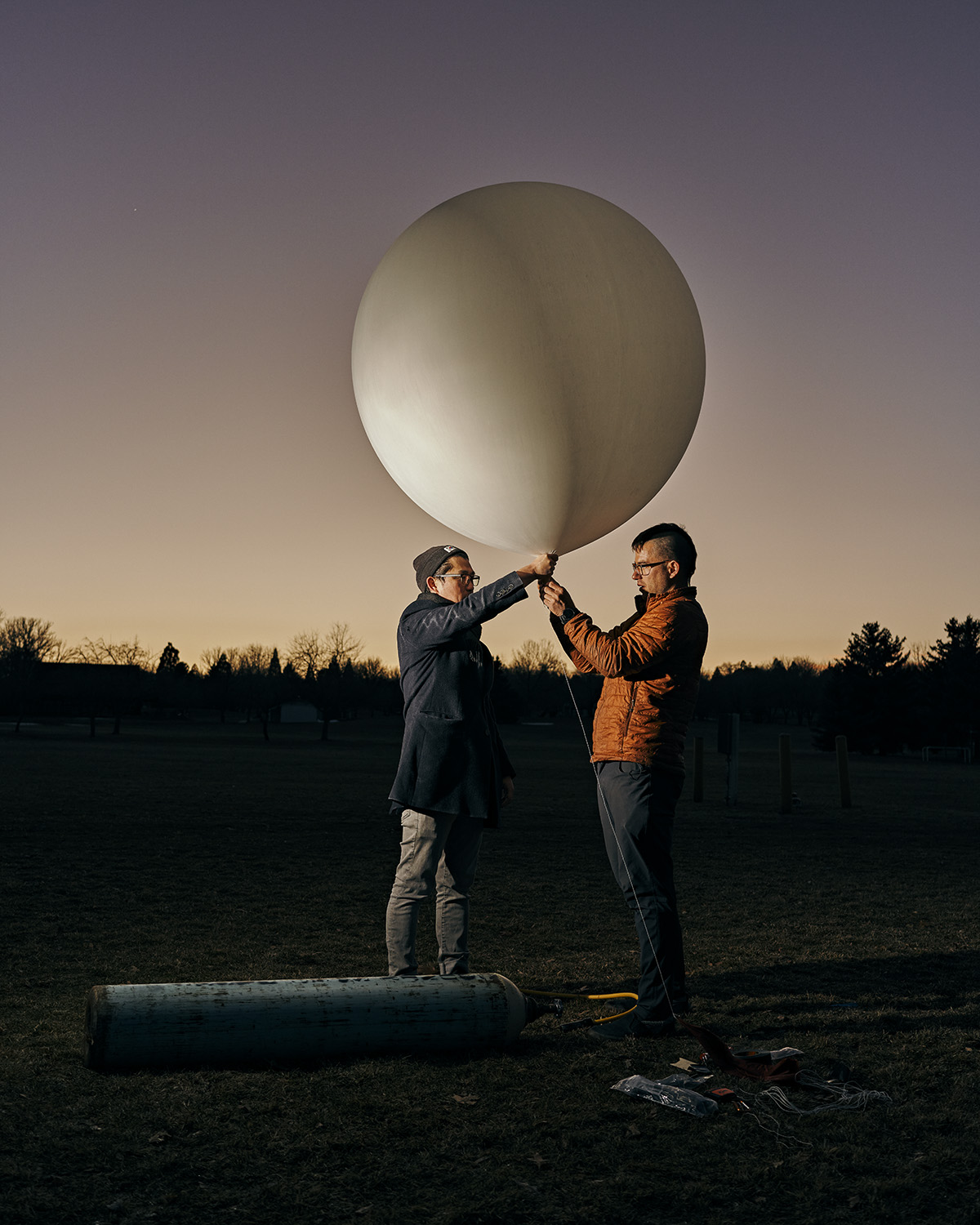Two people outdoors at dusk holding a large white weather balloon, standing on a field of grass, with trees in the distance.