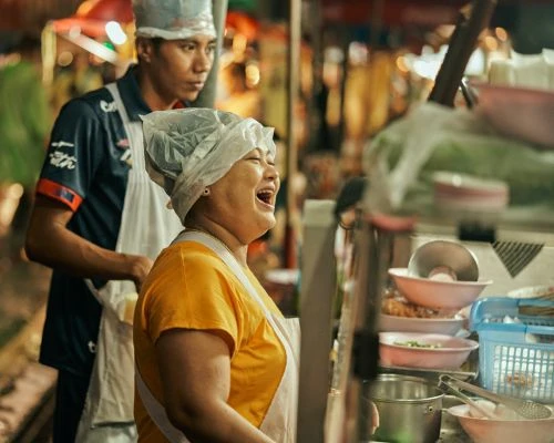 Smiling street food vendor in yellow shirt with plastic cap, with a blurry assistant in the background, prepares dishes outdoors.