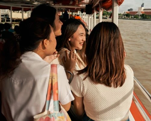 Four women enjoy a boat ride on a river, smiling and looking at the scenery, under an overcast sky with architecture in the distance.
