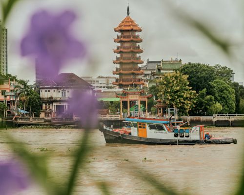 Tugboat navigates a river in front of a tall pagoda in a city with muted colors and foliage in the background on a cloudy day.