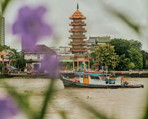 Tugboat navigates a river in front of a tall pagoda in a city with muted colors and foliage in the background on a cloudy day.