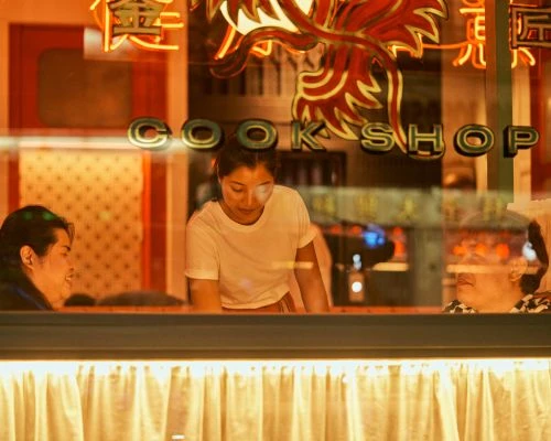 Three people inside a restaurant: one woman standing behind the counter, two people sitting at tables in warm, ambient lighting at night.