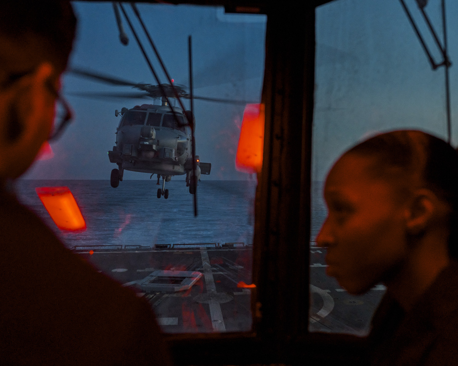 Helicopter approaches a US Navy ship at night, viewed through a window with two silhouetted figures. Dark blue sky above ocean.