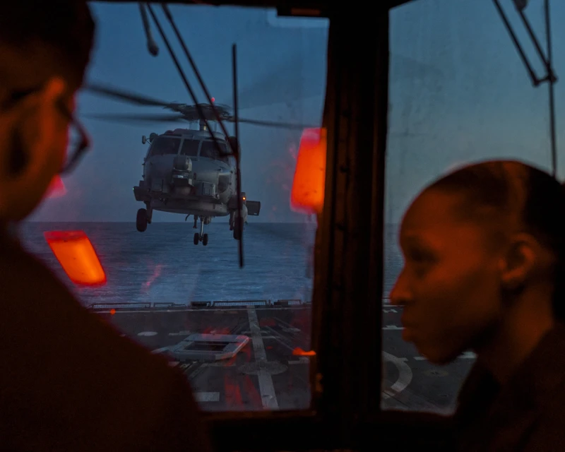 Helicopter approaches a US Navy ship at night, viewed through a window with two silhouetted figures. Dark blue sky above ocean.