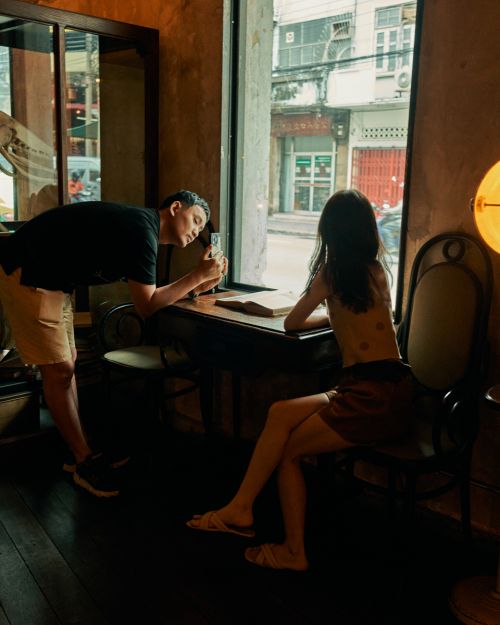 Two people at a table by a window inside a cafe. A man stands and looks out the window while a woman sits and looks at a book.