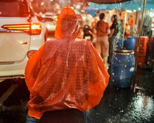 Person in orange raincoat walks in city street during heavy rain with traffic. Evening street scene in urban setting.