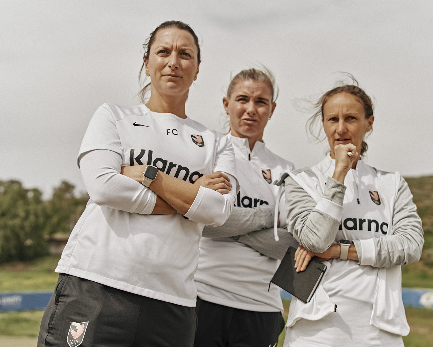 Three adult women in soccer uniforms stand outdoors with arms crossed, featuring athletic wear, smart watches, and a sports environment.