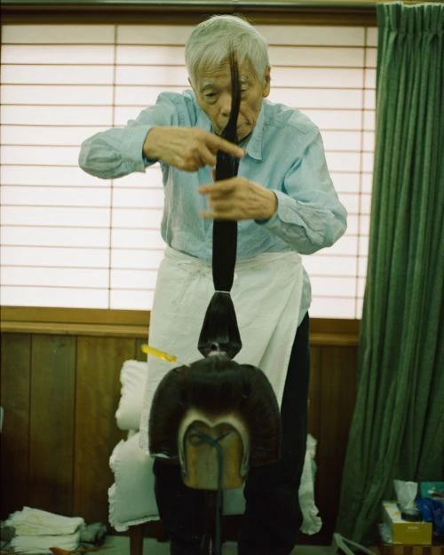 Elderly Asian man in a light-blue shirt and white apron styling a dark wig on a stand in a room with neutral walls and window blinds.