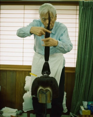 Elderly Asian man in a light-blue shirt and white apron styling a dark wig on a stand in a room with neutral walls and window blinds.