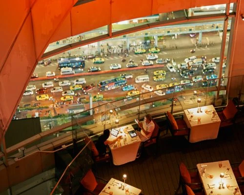 Two adults sit at a restaurant table using tablets, with a bird's-eye view of city streets filled with cars at night beyond a railing.