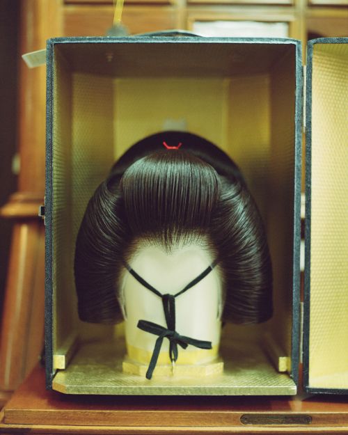 Japanese geisha wig on a mannequin head inside a black box with gold interior, displayed on a wooden surface, studio lighting.