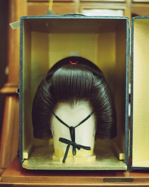 Japanese geisha wig on a mannequin head inside a black box with gold interior, displayed on a wooden surface, studio lighting.