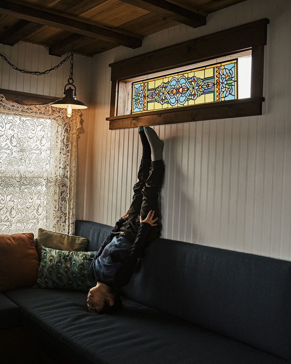 Child lying upside down on a blue couch in a brightly lit room with decorative pillows, a stained-glass window, and warm lighting.