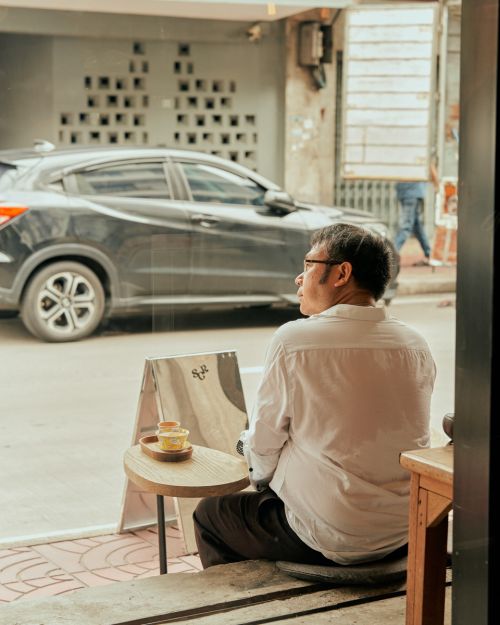 Elderly man in glasses and white shirt sits outside near table with tea and a car parked behind him on city street.