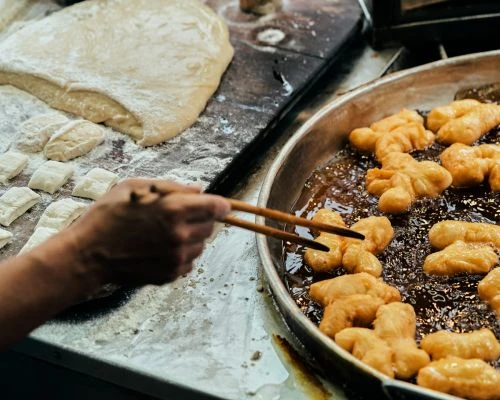 Cooking youtiao (Chinese fried dough) in a metal pan, using chopsticks, next to dough on a floured surface in an indoor setting.