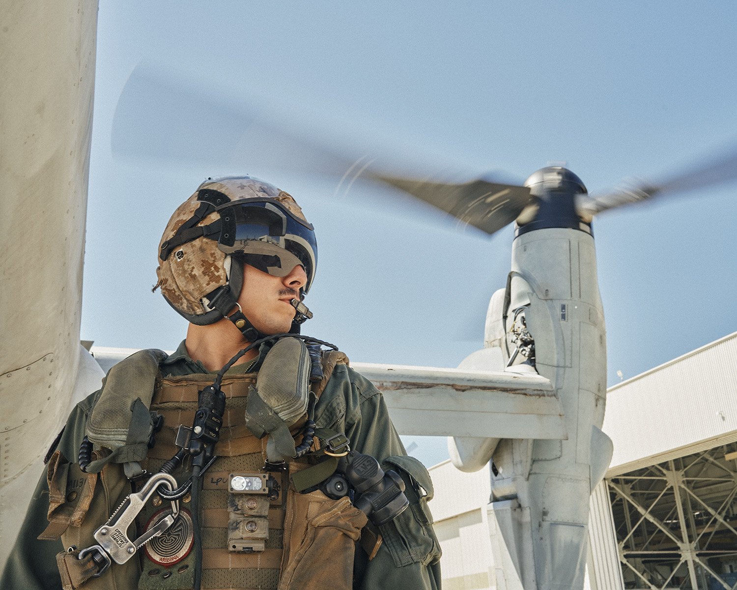 Military personnel in uniform and helmet standing near a tilted rotor aircraft with spinning blades on an outdoor tarmac.