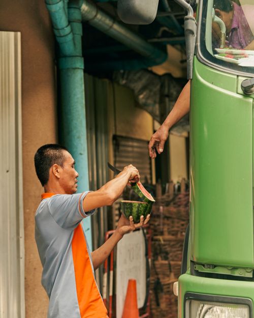 Man in a grey and orange polo shirt holding a halved watermelon next to a green truck, photographed by Balazs Gardi.