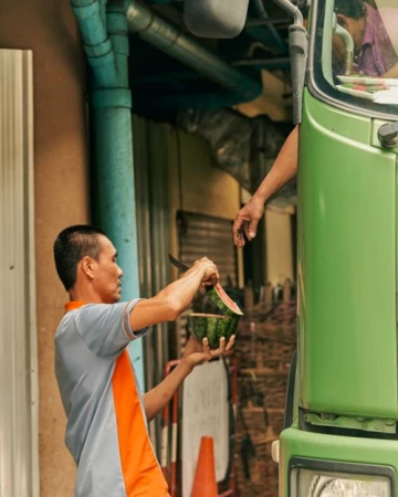 Man in a grey and orange polo shirt holding a halved watermelon next to a green truck, photographed by Balazs Gardi.