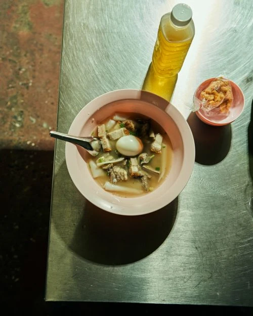Overhead shot of a pink bowl of Asian noodle soup with pork and egg, a bottle of yellow liquid, and cracklings on a metal surface.