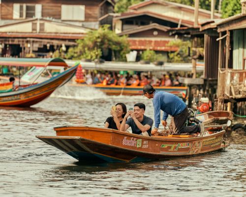 Couple takes photo from longtail boat on Thailand canal waterway, passing waterfront houses and a larger tour boat with passengers.