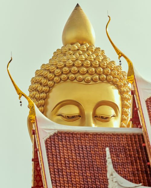 Close-up of a golden Buddha statue with intricate details, viewed from a low angle against a bright sky, partially framed by a temple roof.