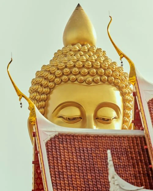 Close-up of a golden Buddha statue with intricate details, viewed from a low angle against a bright sky, partially framed by a temple roof.