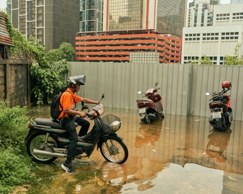 Man in orange shirt on motorcycle in flooded street in Bangkok, Thailand, with buildings and other parked motorcycles in the background.