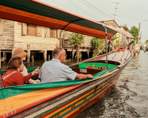 Three people ride in a longtail boat on a river in Thailand. Wooden houses are built on stilts along the riverbank.
