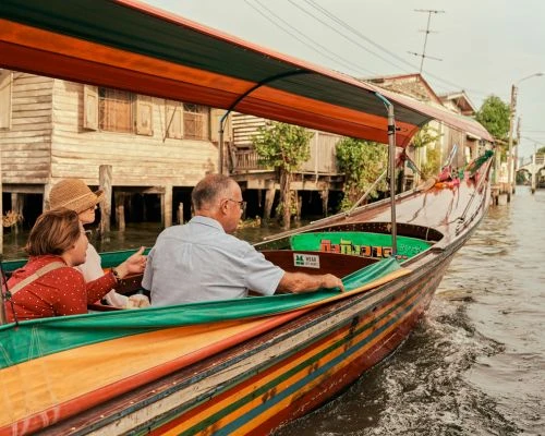 Three people ride in a longtail boat on a river in Thailand. Wooden houses are built on stilts along the riverbank.
