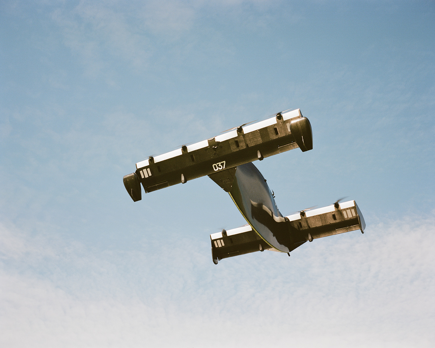 Black and white cargo plane with short wings and tail number 037 flies in clear blue sky on bright sunny day, upward angle shot.