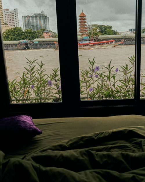 Window view of river in Thailand with boats, a temple, and city buildings during the day, plus a green bed and purple pillow.