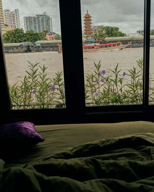 Window view of river in Thailand with boats, a temple, and city buildings during the day, plus a green bed and purple pillow.